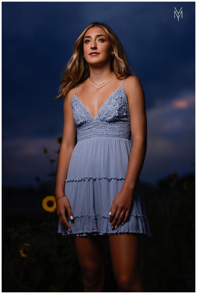 High school senior girl posing in front of sunflowers in a blue sundress.