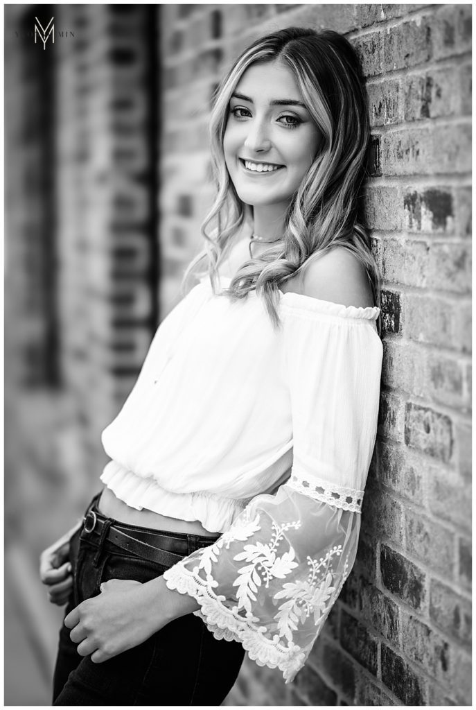 Black and white image of a high school senior girl leaning against a wall for her senior pictures.