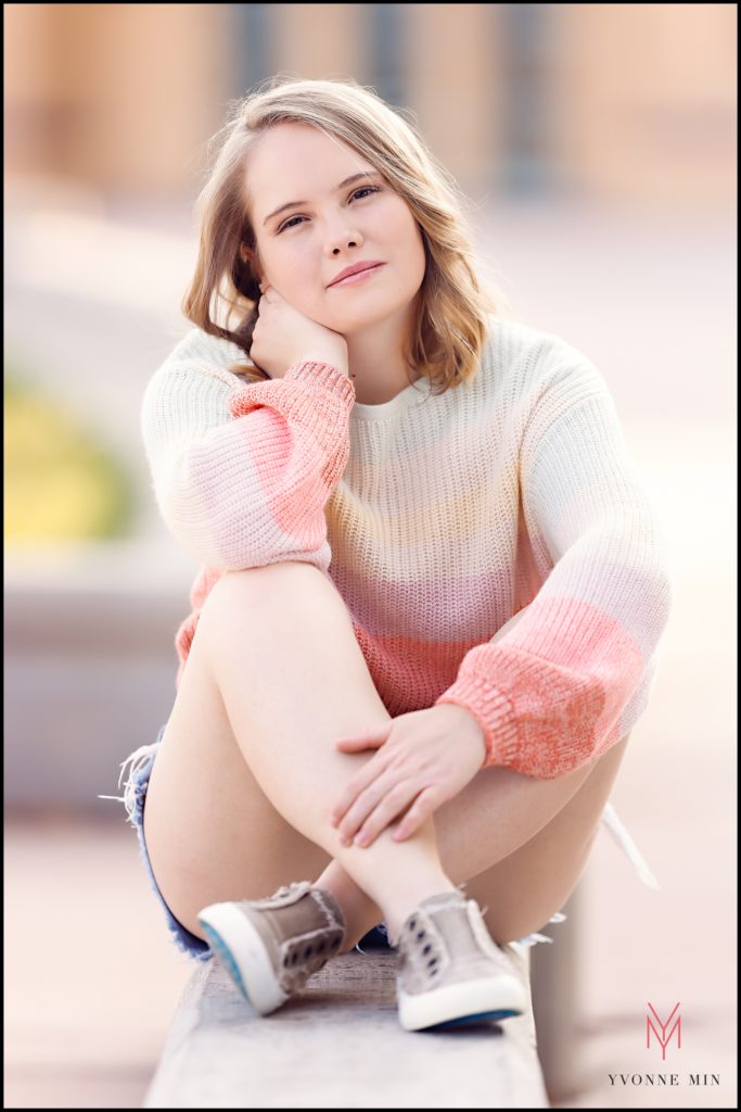 A high school senior girl sits on the concrete ledge which surrounds the lake at City Park in Denver, Colorado.