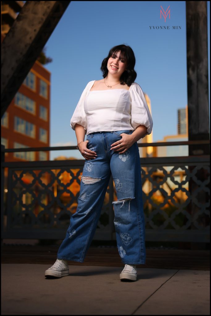 Sammy poses for her senior photos on a bridge in LoDo with downtown buildings behind her.