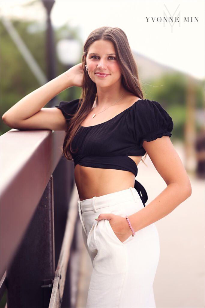A senior girl poses in a black shirt and white pants on a bridge in downtown Golden, Colorado with Yvonne Min Photography.