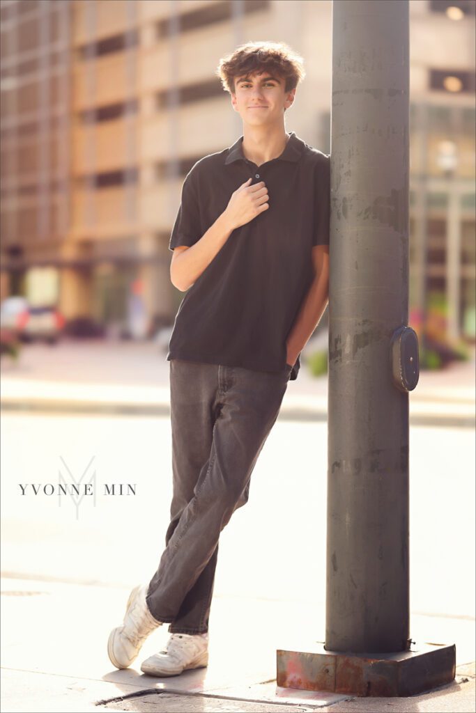A high school senior boy leans against a traffic light post during his senior photoshoot with Yvonne Min Photography in downtown Denver.