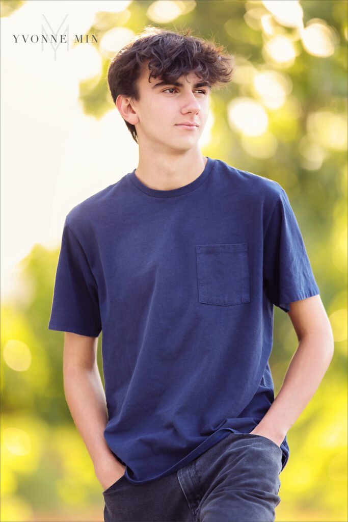 A high school senior boy poses in front of trees in a blue shirt during his senior photoshoot with Yvonne Min Photography in Denver.