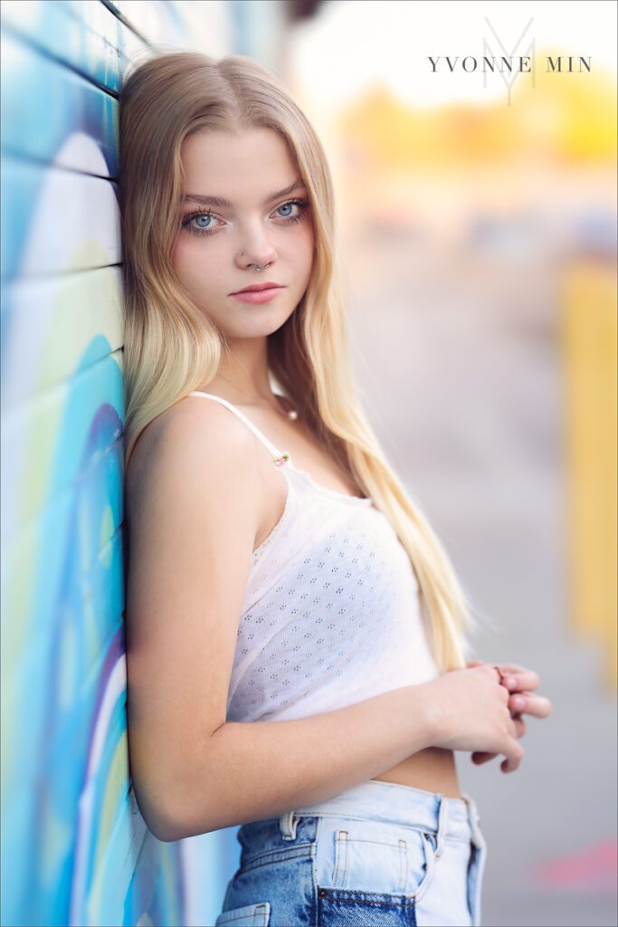 A high school senior girl poses in RiNo in Downtown Denver against a wall while being photographed by Yvonne Min Photography.