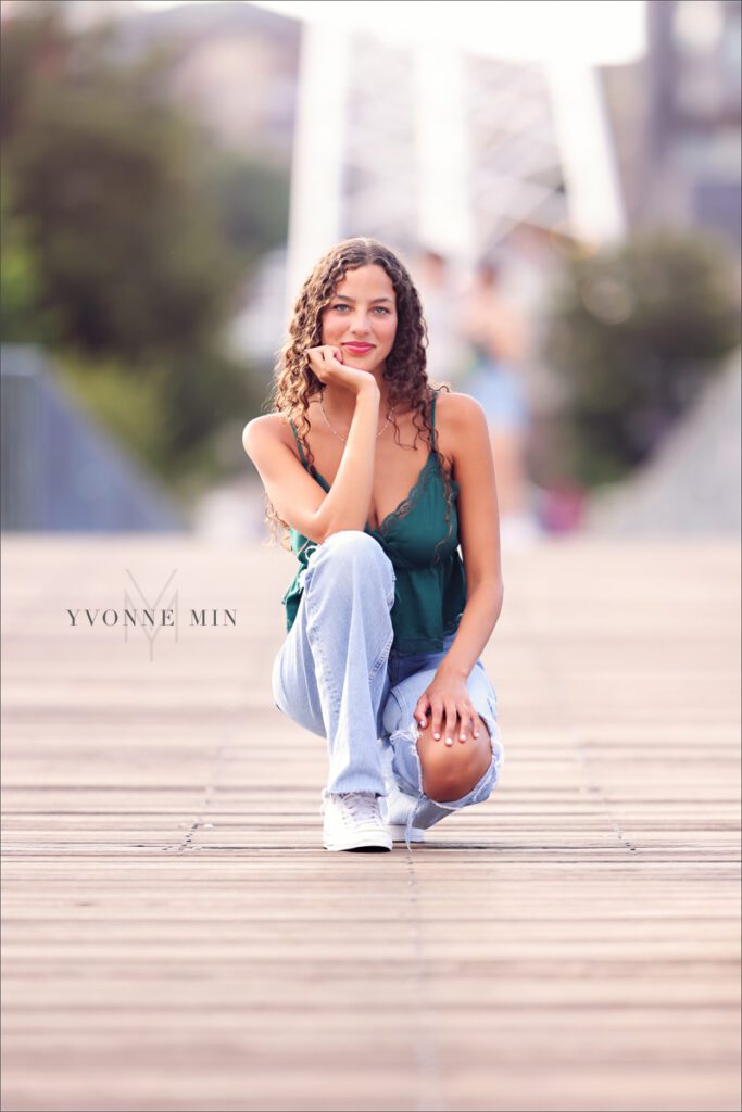 A high school senior squats on a bridge at her senior photoshoot in downtown Denver with Yvonne Min Photography.