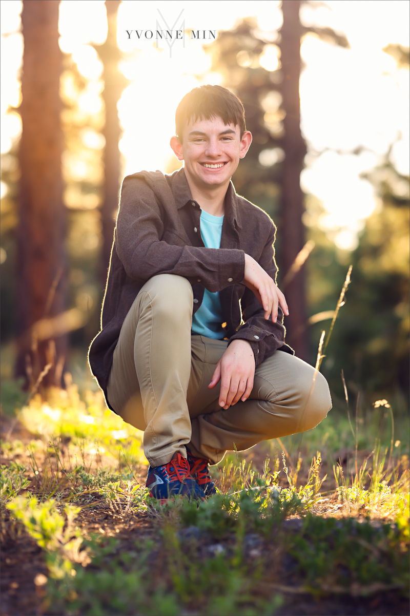 A senior boy from Stargate High School poses in a squat on a trail at Chautauqua Park in Boulder, Colorado in a brown jacket with Yvonne Min Photography.