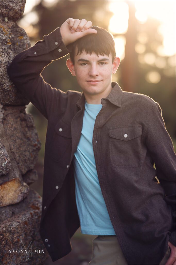 A senior boy from Stargate High School leans against a stone wall on a trail at Chautauqua Park in Boulder, Colorado in a brown jacket with Yvonne Min Photography.