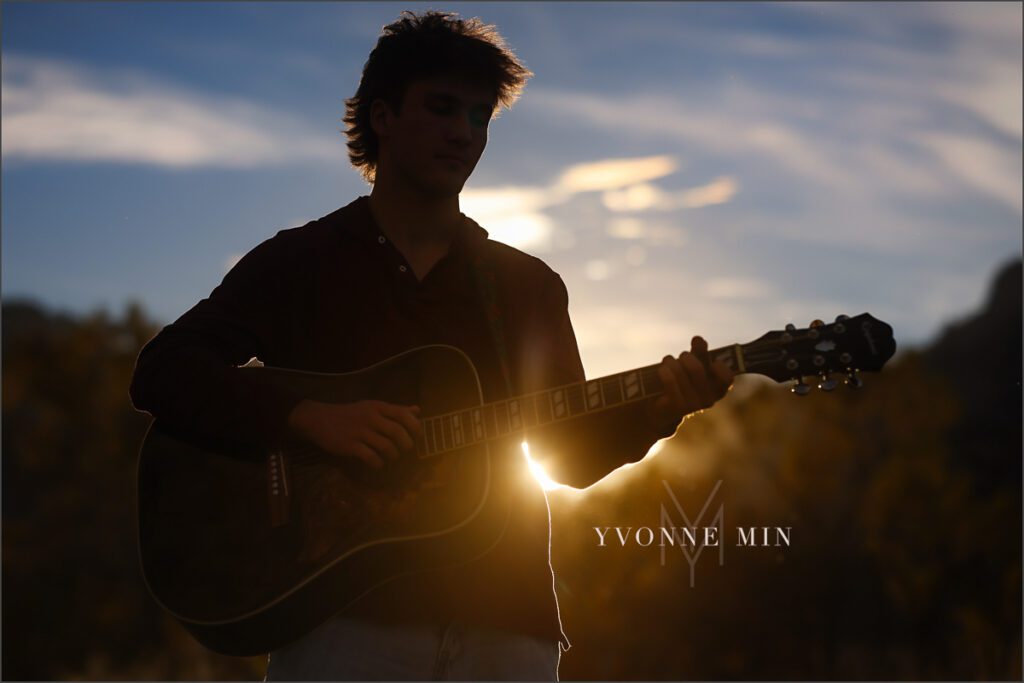 A senior guy from Nothfield High Schools plays a guitar during his senior photoshoot at South Mesa Trailhead outside Boulder with Yvonne Min Photography. This photograph is a silhouette with the sun peaking out behind him.