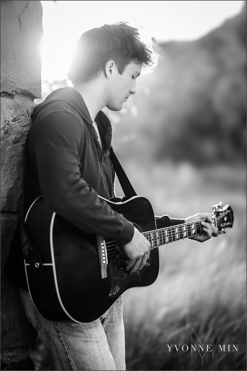 A black and white senior photo of a guy from Northfield High School leaning on a wall playing a guitar at South Mesa, Boulder taken by Yvonne Min Photography.