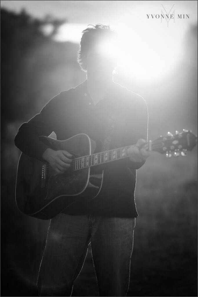 A senior guy from Nothfield High Schools plays a guitar during his senior photoshoot at South Mesa Trailhead outside Boulder with Yvonne Min Photography. This photograph is a silhouette with the sun peaking out behind him.