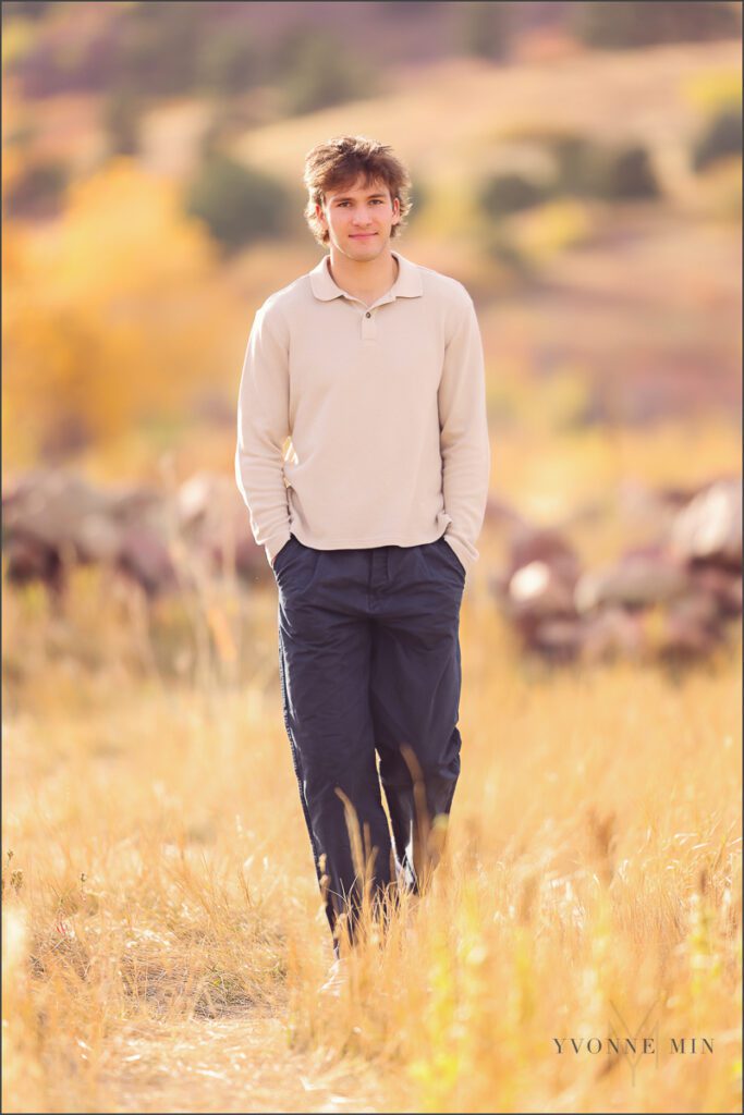 A senior boy wearing a cream sweater walks in a field during his senior photoshoot at South Mesa in Boulder with Yvonne Min Photography.